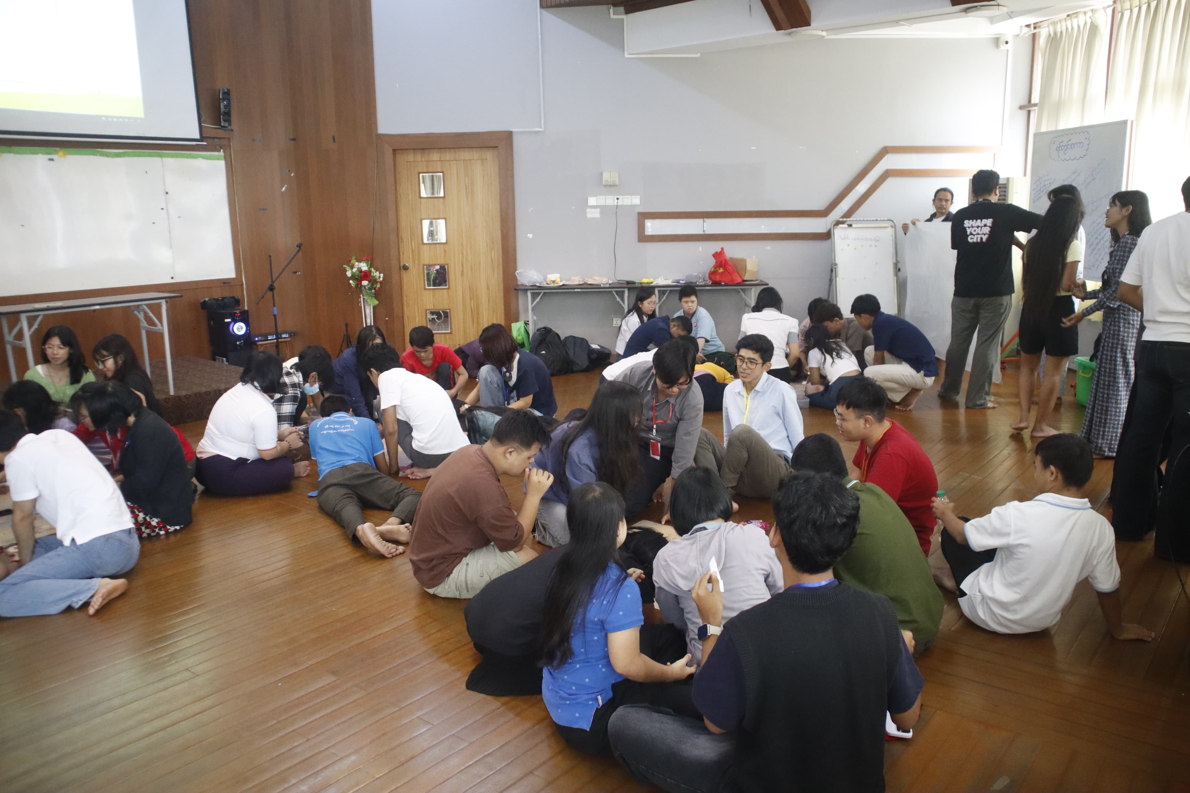 Participants sitting in groups on the wooden floor of the workshop hall